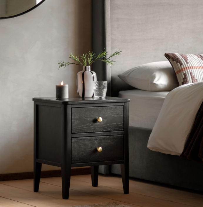 Gallery Direct Acton 2 Drawer Bedside in Black with gold knob handles next to bed and on oak wood floor with vase & flowers