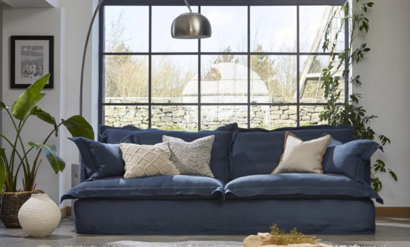 Tetrad Ava Grand Sofa in denim blue linen in a modern industrial living room with large windows and slouchy feather cushions.